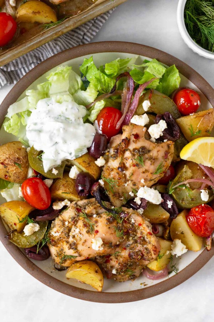 Plate filled with roasted chicken thighs, roasted potatoes, peppers, and onions, cherry tomatoes, olives, feta, romaine, and tzatziki sauce. Next to the plate is a dish of dill and a pan full of veggies off to the side.