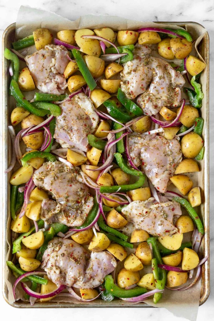 Baking sheet with raw chicken thighs, quartered potatoes, sliced green peppers, and sliced red onion all covered in oil and spices.