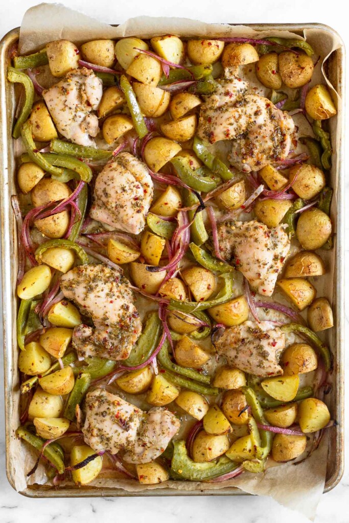 Baking sheet with seasoned baked chicken thighs, roasted quartered potatoes, roasted sliced green peppers, and roasted sliced red onion.