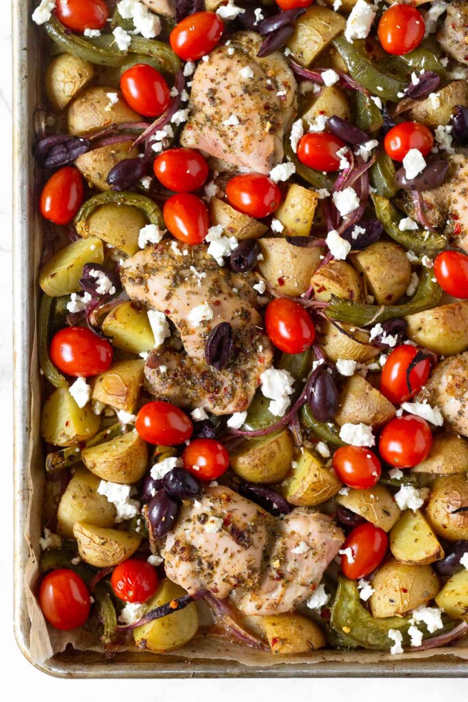 Baking sheet filled with roasted potatoes, green peppers, red onion, tomatoes, olives, and feta with roasted chicken thighs nestled in-between everything.