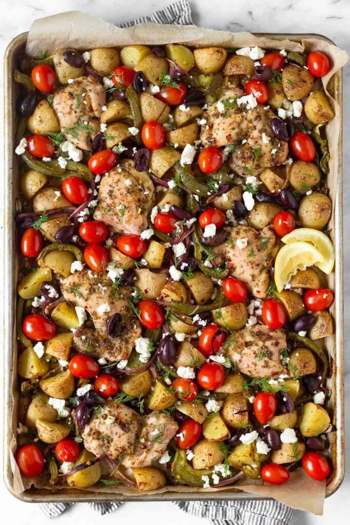 Mediterranean sheet pan chicken with potatoes, peppers, onions, cherry tomatoes, Kalamata olives, and feta. It is garnished two lemon wedges and fresh dill.