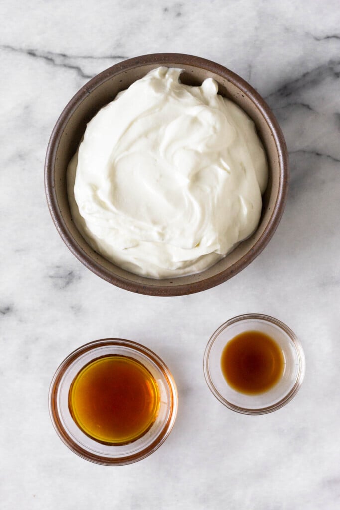 White marble counter with a bowl of greek yogurt, a bowl of vanilla extract, and a bowl of maple syrup.