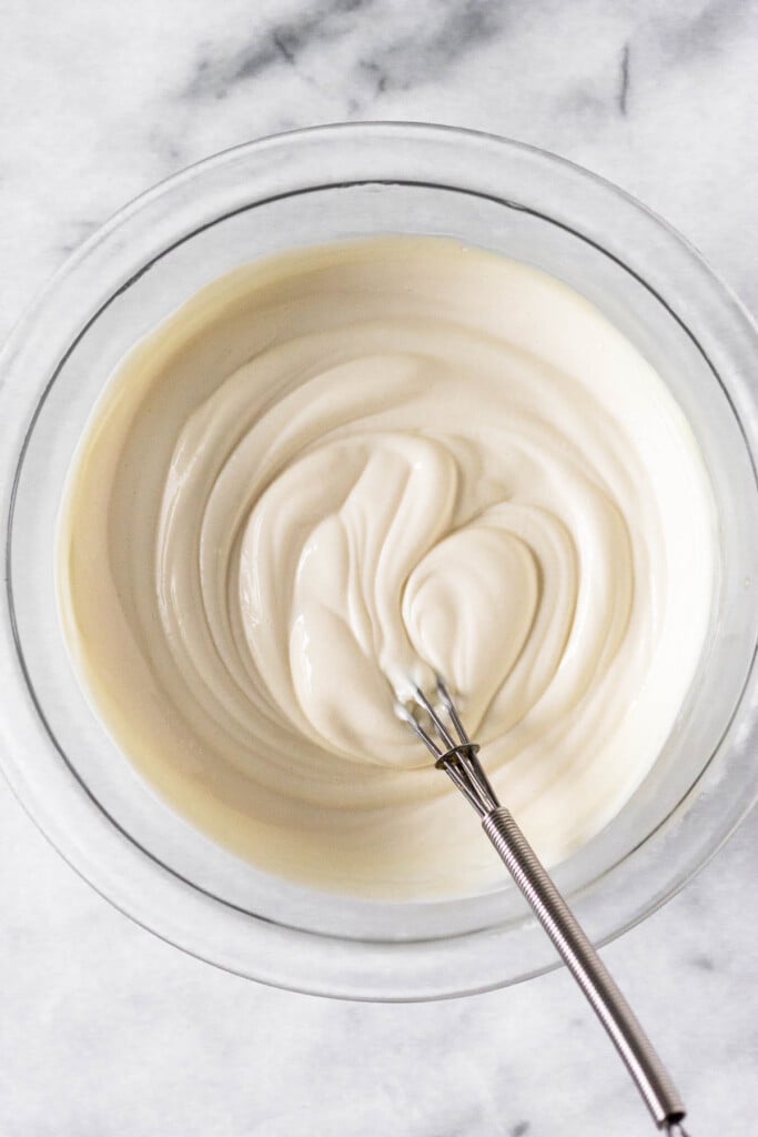 A glass bowl of greek yogurt frosting with a small metal whisk in it.