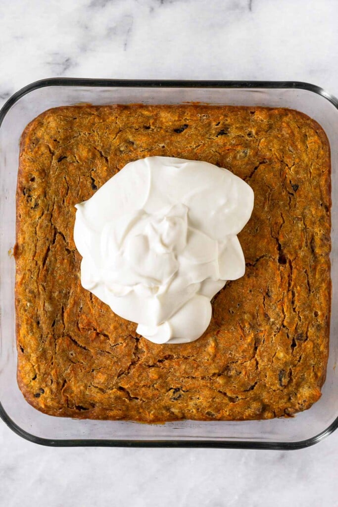 Healthy carrot cake bars in a glass baking dish with a pound of greek yogurt icing on top of them before it is spread.