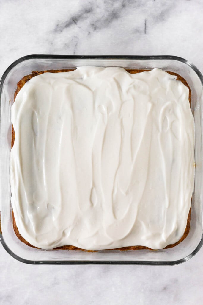 Frosting with greek yogurt spread across cake in a square baking dish.