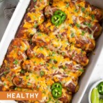 Healthy chicken enchilada with red sauce and cheese in a large casserole dish. They are garnished with sliced jalapeños and cilantro. Around the dish is a bowl of lime wedges, a bowl of cilantro, and a bowl of tortilla chips.