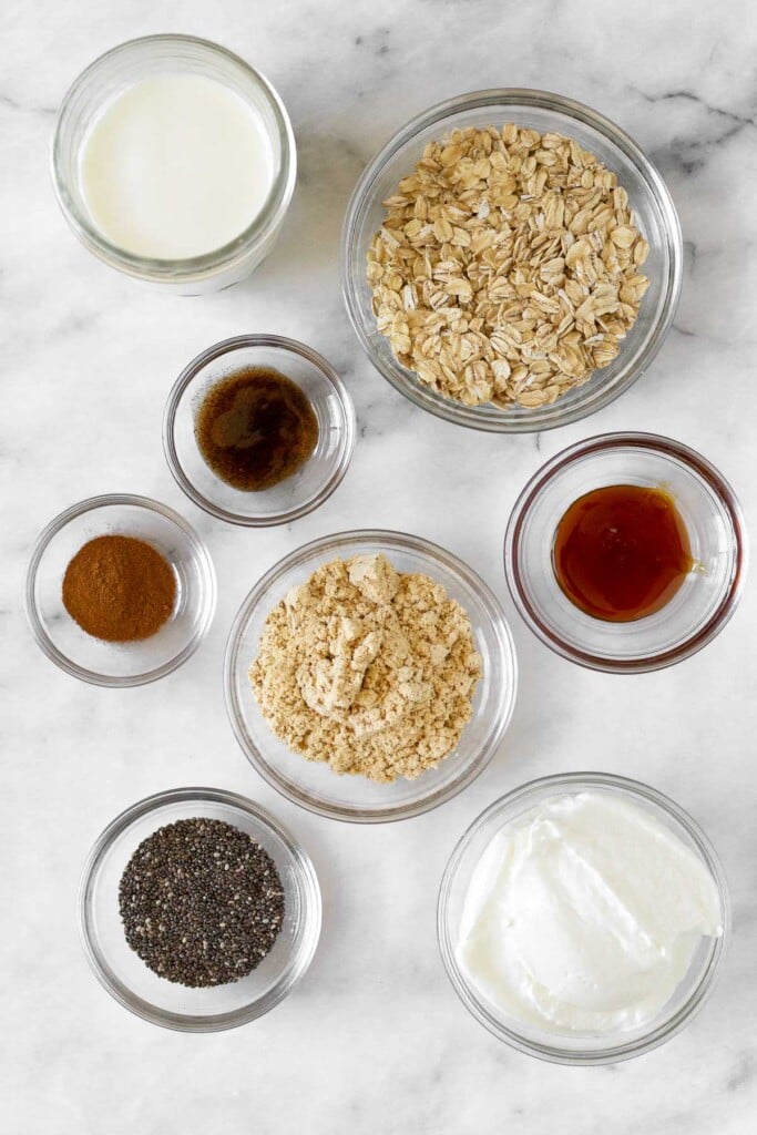 White marble counter with a bowl of rolled oats, a bowl of honey, a bowl of greek yogurt, a bowl of chia seeds, a bowl of vanilla protein powder, a bowl of cinnamon, a bowl of vanilla extract, and a jar of milk.