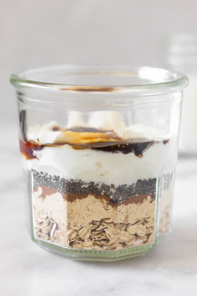 A straight on shot of a jar layered with rolled oats, protein powder, cinnamon, chia seeds, greek yogurt, honey, and vanilla extract. Behind it is a jar of milk.