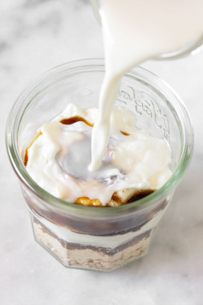 A straight on shot of a jar layered with rolled oats, protein powder, cinnamon, chia seeds, greek yogurt, honey, and vanilla extract with milk being poured into it.