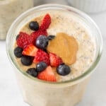 A jar of protein overnight oats topped with fresh diced strawberries, blueberries, and peanut butter. Around it is another jar of overnight oats, a small dish of berries, and a teaspoon of cinnamon.