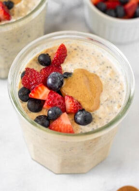 A jar of protein overnight oats topped with fresh diced strawberries, blueberries, and peanut butter. Around it is another jar of overnight oats, a small dish of berries, and a teaspoon of cinnamon.