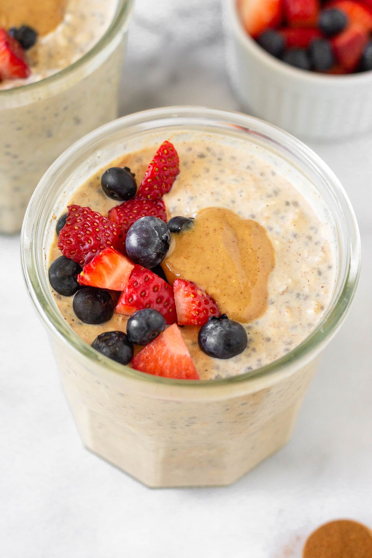 A jar of protein overnight oats topped with fresh diced strawberries, blueberries, and peanut butter. Around it is another jar of overnight oats, a small dish of berries, and a teaspoon of cinnamon.