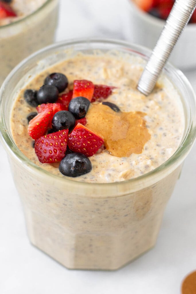 A jar of high protein overnight oats topped with strawberries, blueberries, and peanut butter with a spoon in the jar. Behind it is another jar of overnight oats and a small ramekin of fresh berries.