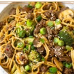 Beef and broccoli noodles in a white bowl garnished with sliced green onions and sesame seeds.