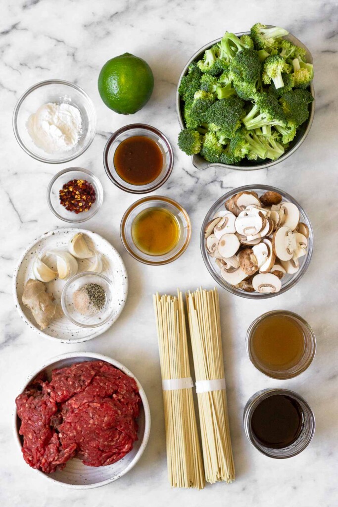 White marble counter with a bowl of broccoli florets, a bowl of sliced mushrooms, a jar of bone broth, a jar of coconut aminos, two bundles of ramen noodles, a bowl of raw ground beef, a plate of fresh ginger, 4 garlic cloves, and a small bowl of salt and pepper, a bowl of honey, a bowl of sesame oil, a bowl of red pepper flakes, a bowl of arrowroot flour, and a whole lime.