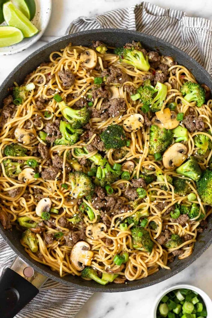 Beef and broccoli noodles with mushrooms in a large skillet garnished with sliced green onions and sesame seeds. The pan is sitting on a striped towel and next to it is a plate of lime wedges and a bowl of green onions.
