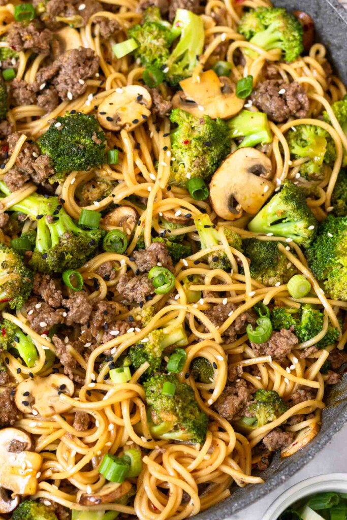 Ramen noodles with beef, broccoli, and mushrooms tossed with stir fry sauce in a large skillet. Everything is garnished with sliced green onions and sesame seeds.