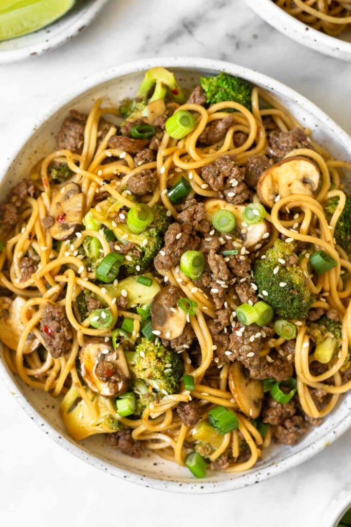 Overhead shot of ramen noodle stir fry with beef, broccoli, and mushrooms. It is garnished with sliced green onions and sesame seeds.