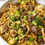 Beef and broccoli noodles in a white bowl garnished with sliced green onions and sesame seeds. Behind the bowl is a plate of lime wedges and another bowl of noodles.