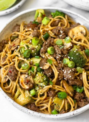 Beef and broccoli noodles in a white bowl garnished with sliced green onions and sesame seeds. Behind the bowl is a plate of lime wedges and another bowl of noodles.