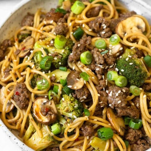Beef and broccoli noodles in a white bowl garnished with sliced green onions and sesame seeds. Behind the bowl is a plate of lime wedges and another bowl of noodles.