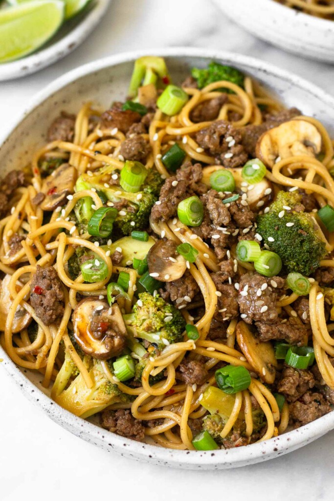 Beef and broccoli noodles in a white bowl garnished with sliced green onions and sesame seeds. Behind the bowl is a plate of lime wedges and another bowl of noodles.