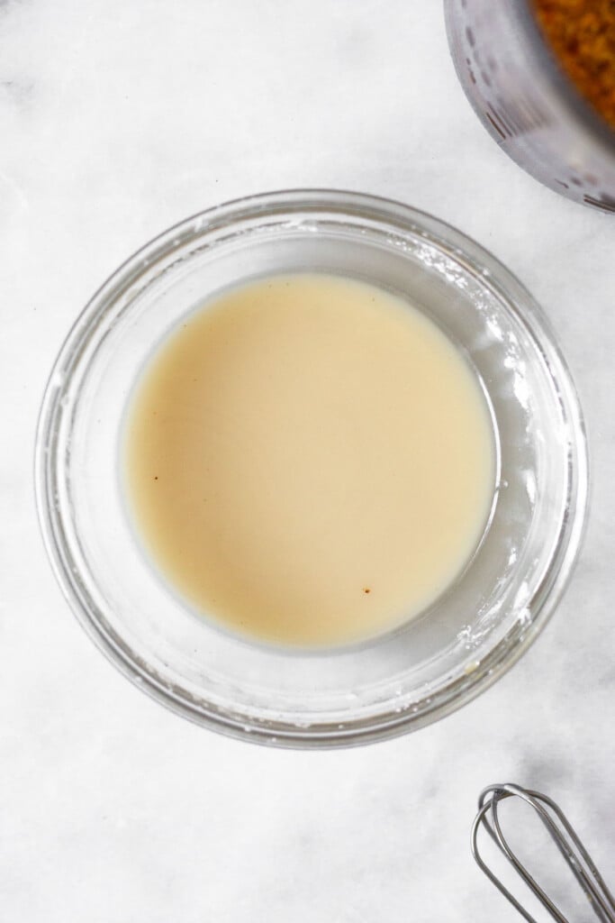 Small bowl filled with a slurry of water and arrowroot flour. Next to it is a small whisk and a jar filled with stir fry sauce.