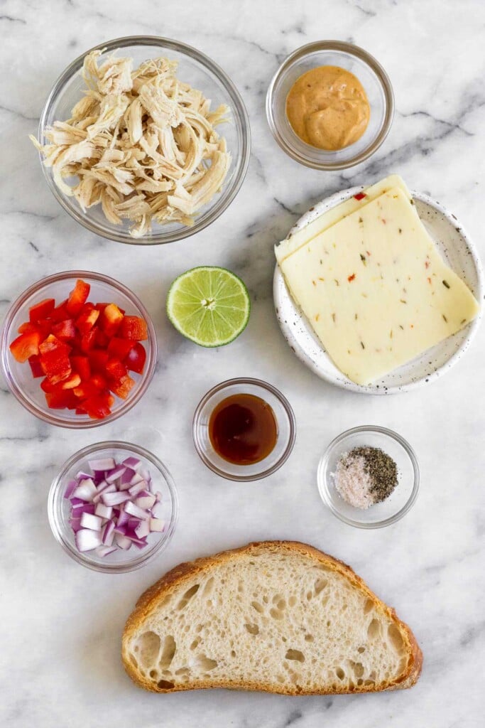 White marble counter with a bowl of chipotle mayo, a plate of 2 slices of pepper jack cheese, a bowl of salt and pepper, a big slice of sourdough bread, a bowl of honey, a bowl of diced red onion, a bowl of diced red bell pepper, and a bowl of shredded chicken.