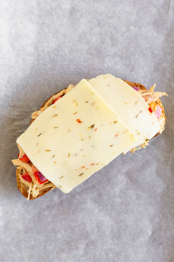 A piece of toasted sourdough bread topped with chicken salad and 2 pieces of sliced cheese on a piece of parchment paper.