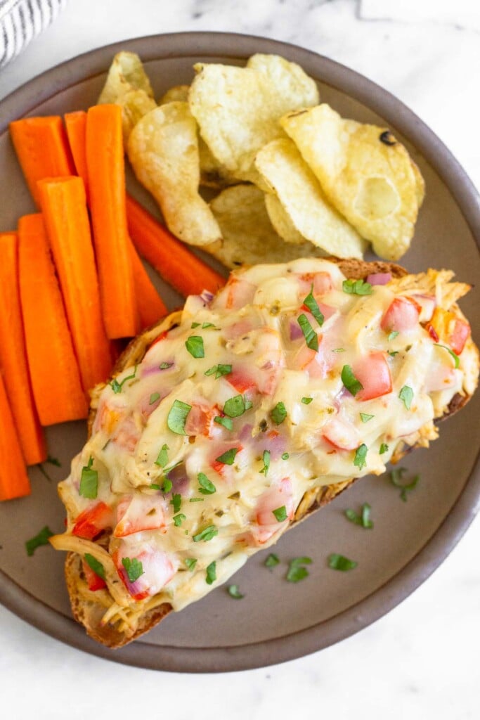 Overhead shot of a brown plate with a chicken salad melt garnished with cilantro, carrot sticks, and potato chips on it.