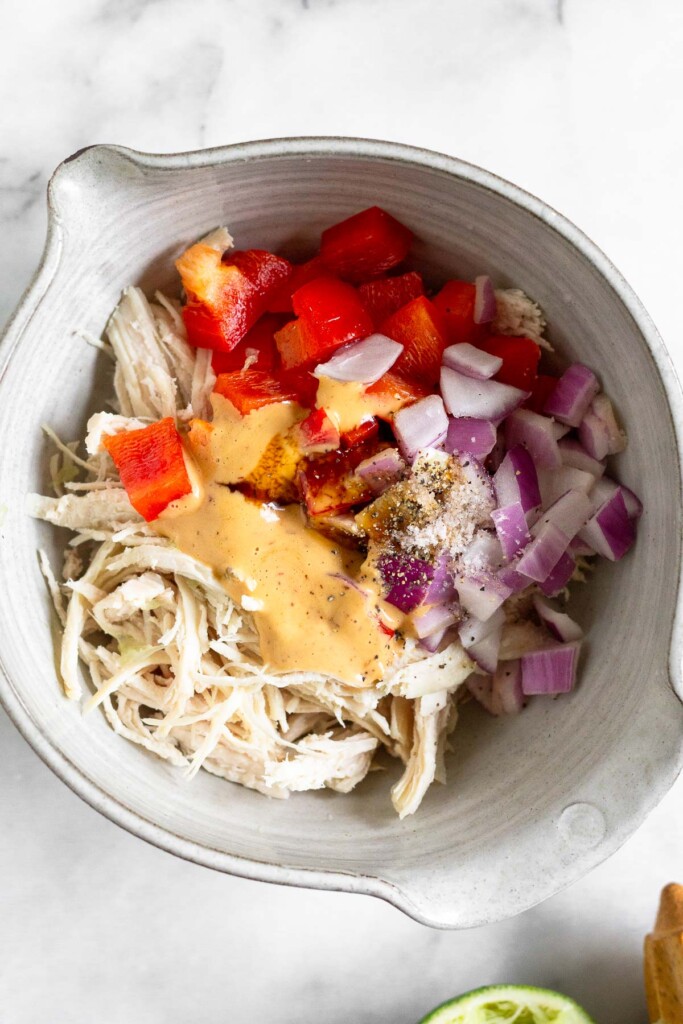 Bowl of shredded chicken, diced red bell pepper, diced red onion, chipotle mayo, honey, salt, and pepper before it is mixed together. Next to the bowl is half a juiced lime and a wooden reamer.