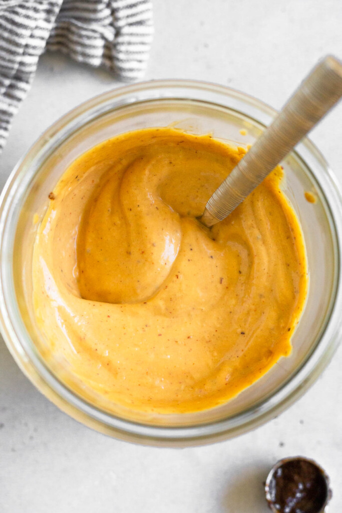 A overhead shot jar filled with homemade chipotle mayo with a spoon it it. Next to the jar is a striped towel and a teaspoon with chipotle paste in it.