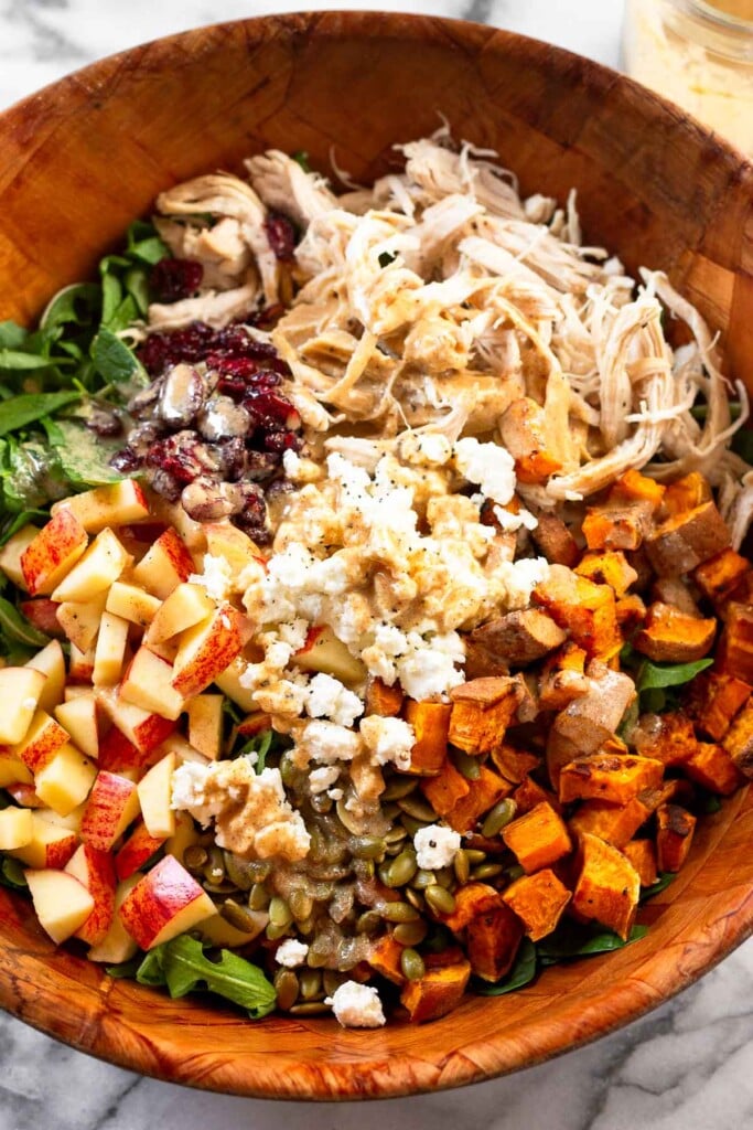 Large wooden salad bowl with shredded chicken, roasted sweet potatoes, pepitas, diced apple, dried cranberries, and goat cheese over mixed greens with dressing on it.