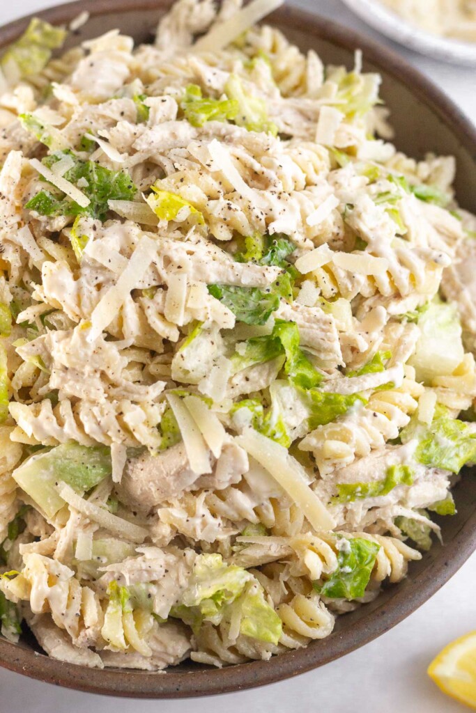 A large bowl of chicken caesar pasta salad. Next to it is half a juiced lemon and behind it is a bowl of grated parmesan cheese.