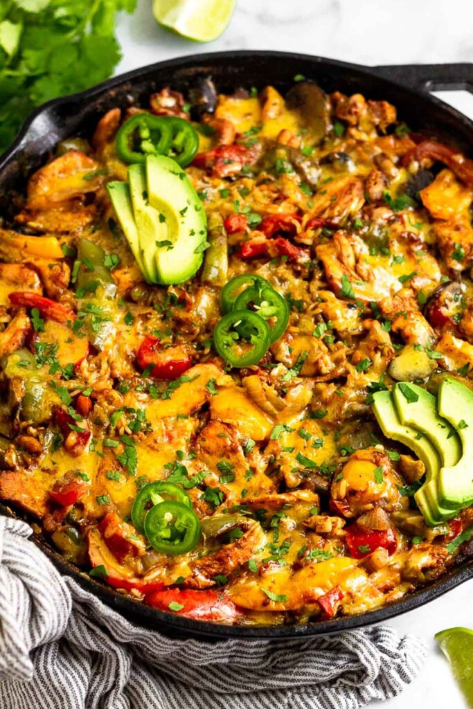 High protein chicken casserole with rice, bell peppers, and melted cheese in a large cast iron pan. It is garnished with sliced avocado, sliced jalapeño, and cilantro. Around the pan is more cilantro and lime wedges.