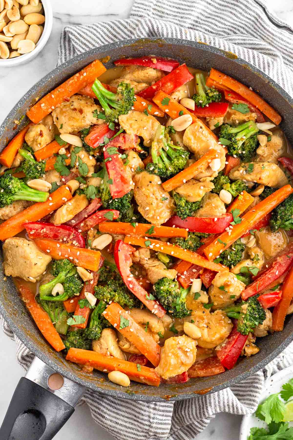 High protein chicken recipe in a skillet with chicken breasts, bell peppers, and broccoli tossed in a peanut sauce. It is topped with sesame seeds, peanuts, and fresh cilantro. The pan is sitting on a striped towel with a bowl of peanuts next to it and a plate of cilantro.