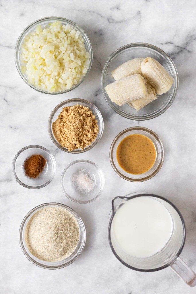 White marble counter with a bowl of frozen cauliflower rice, a bowl of frozen banana pieces, a bowl of peanut butter, a jar of milk, a bowl of protein powder, a bowl of salt, a bowl of cinnamon, and a bowl of peanut butter powder.