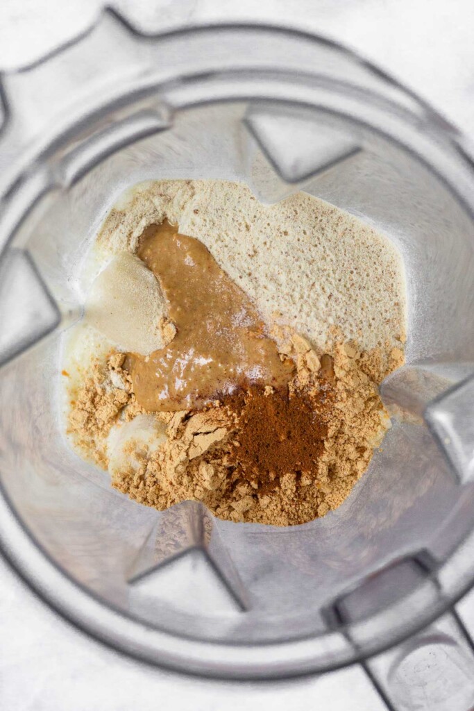Overhead shot of a blender with frozen banana pieces, protein powder, peanut butter powder, peanut butter, and cinnamon.