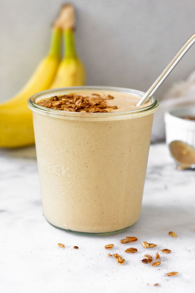 Banana peanut butter protein smoothie in a jar topped with granola and a metal straw in it. In front of it are pieces of granola and behind it are two bananas, a small dish of granola, and a spoon with peanut butter on it.