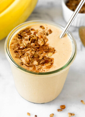Peanut butter banana protein smoothie in a glass jar topped with granola and cinnamon with a straw in it. Around the jar are some bananas and a bowl of granola.