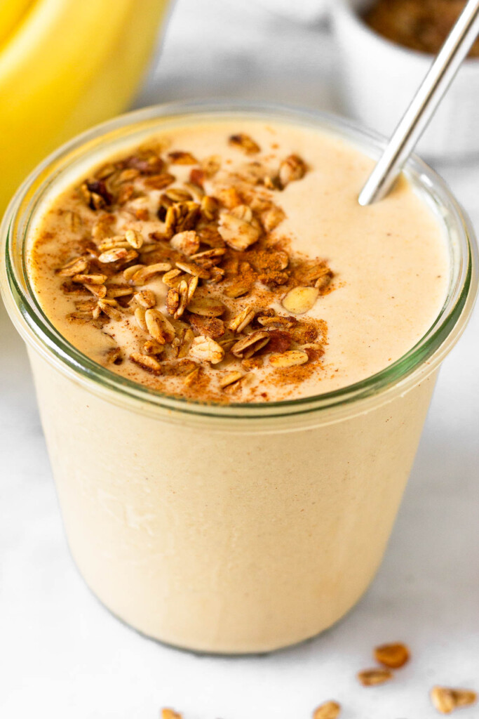 Peanut butter protein smoothie in a jar. It is topped with granola and cinnamon and a metal straw is in the jar. Behind the jar is a bunch of banana and a small dish of granola.