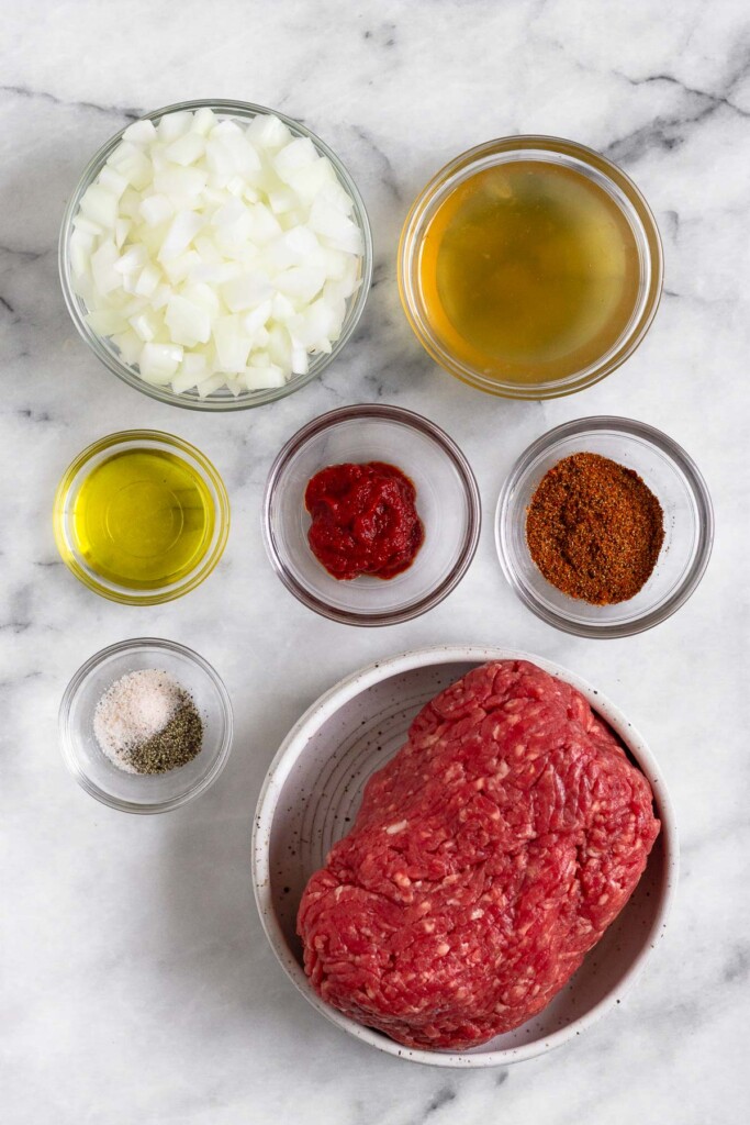 White marble counter with a bowl of chicken broth, a bowl of taco seasoning, a bowl of raw round beef, a bowl of salt and pepper, a bowl of olive oil, a bowl of tomato paste, and a bow of chopped onions.
