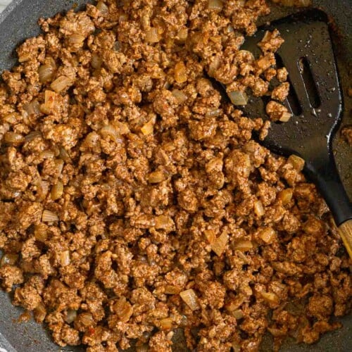 Ground beef taco meat in a large skillet with a spatula in it. The pan is sitting on a striped towel.