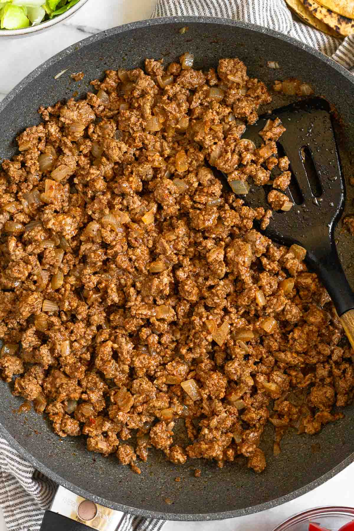 Ground beef taco meat in a large skillet with a spatula in it. The pan is sitting on a striped towel.