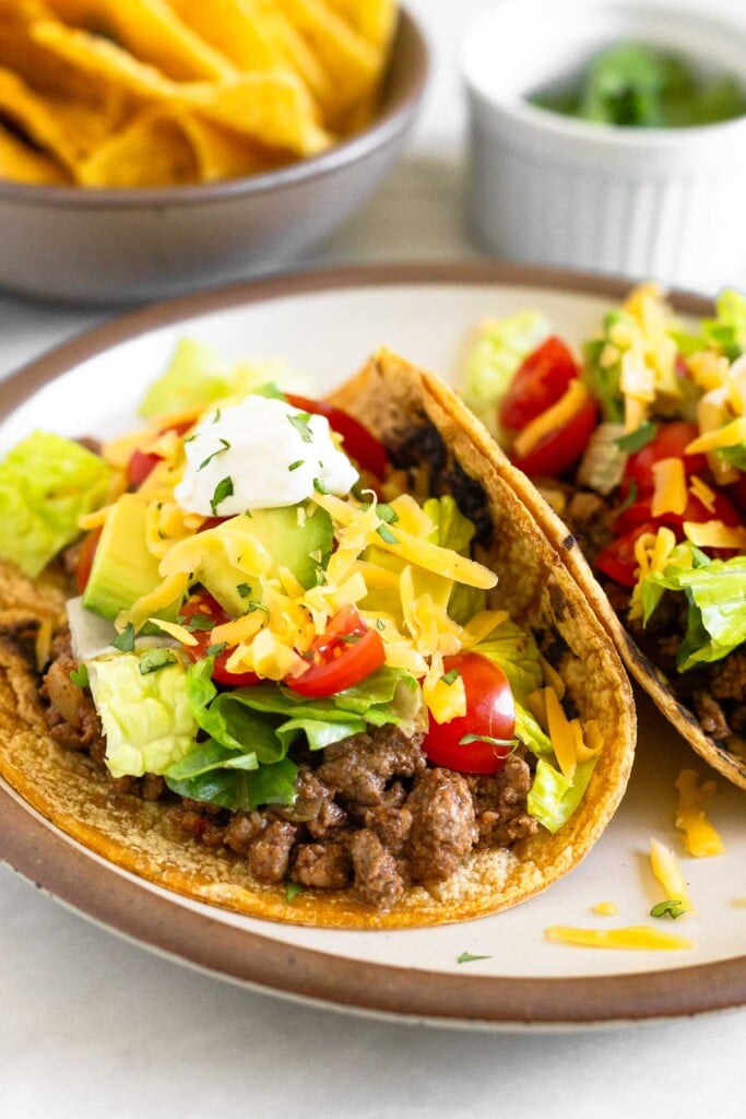 A close-up of a plate with two tacos on it. They have ground beef, lettuce, tomato, cheese, avocado, sour cream, and cilantro on corn tortillas. Behind the plate is a bowl of tortilla chips and a bowl of cilantro.