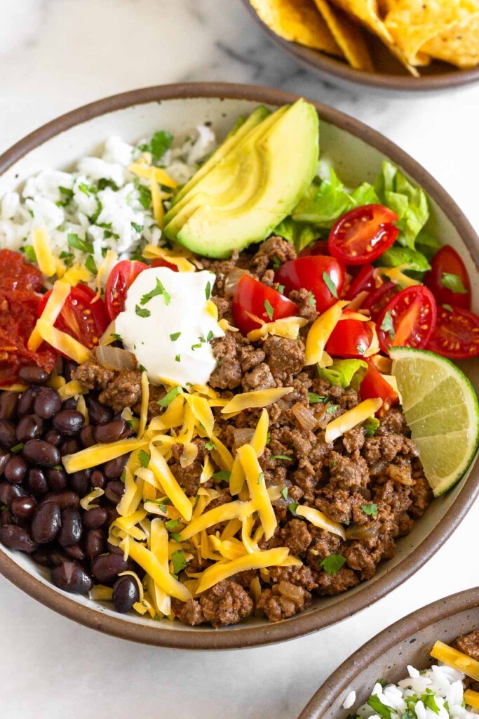 A large white bowl filled with ground beef taco meat, black beans, salsa, cilantro lime rice, slice avocado, lettuce, tomatoes, shredded cheese, sour cream, and a lime wedge. It is garnished with cilantro. In front of the bowl is another taco bowl and behind it is a bowl of tortilla chips.