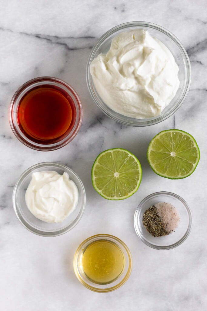 White marble counter with a bowl of greek yogurt, a lime cut in half, a bowl of salt and pepper, a bowl of honey, a bowl of sour cream, and a bowl of red wine vinegar.