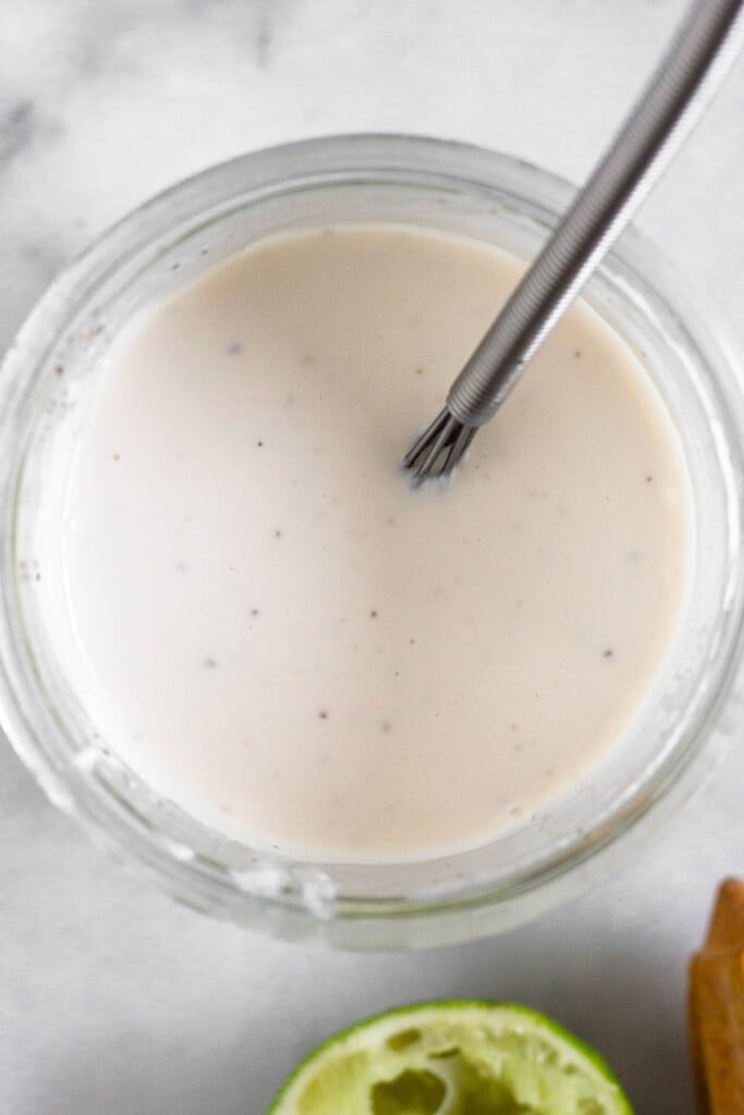 Overhead shot of a jar of healthy coleslaw dressing with a small whisk in it. Next to the jar is half a juiced lime and a wooden reamer.