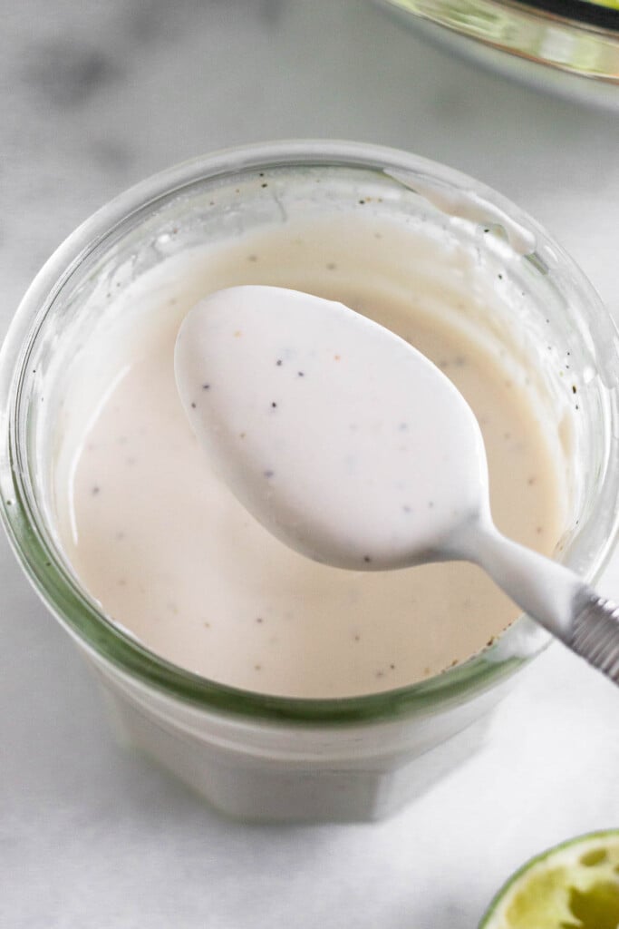 Greek yogurt coleslaw dressing in a jar with a spoon full of dressing overtop of it. In front of the jar is half a juiced lime.