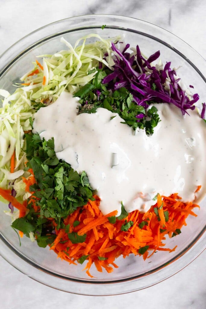 Glass bowl with piles of shredded green cabbage, shredded purple cabbage, shredded carrots, and cilantro with healthy coleslaw dressing poured overtop of it.